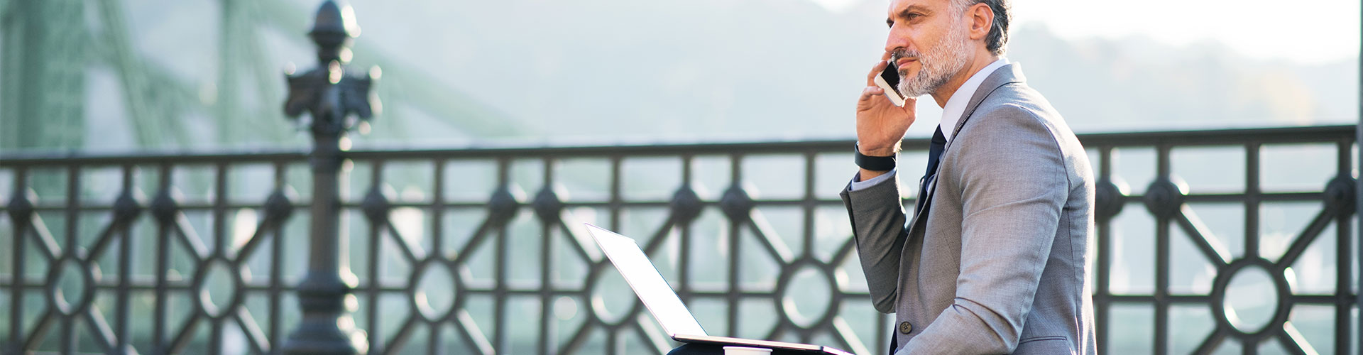 Fondos de Inversion - Hombre maduro de negocios con traje gris, hablando por el movil y portatil, sentado en un banco en una ciudad
