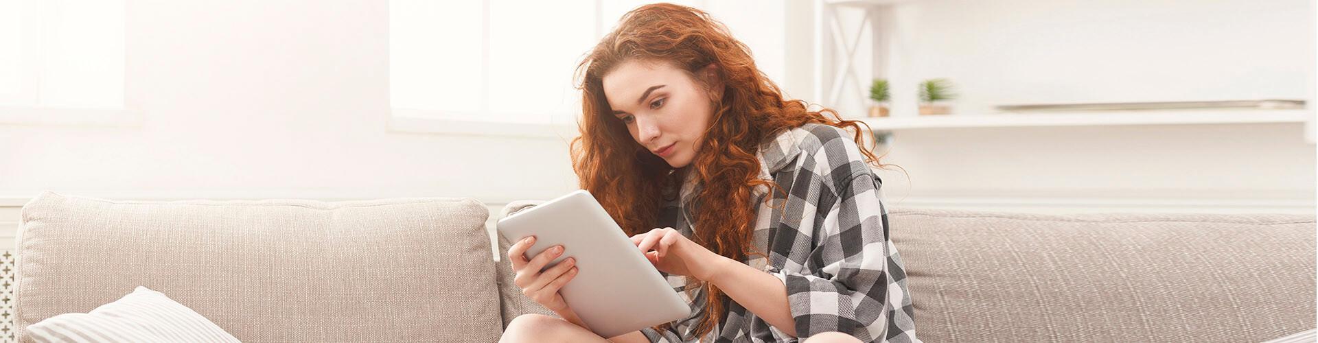 Cuenta de Pago Básica - Chica joven con una camisa de cuadros sentada en el sofá de su casa con una tablet buscando cuentas